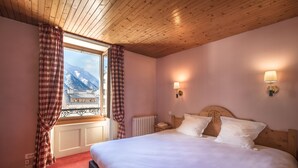 Un dormitorio con techo de madera, una cama grande con ropa blanca, vistas a montañas a través de una ventana y dos lámparas fijadas a la pared.