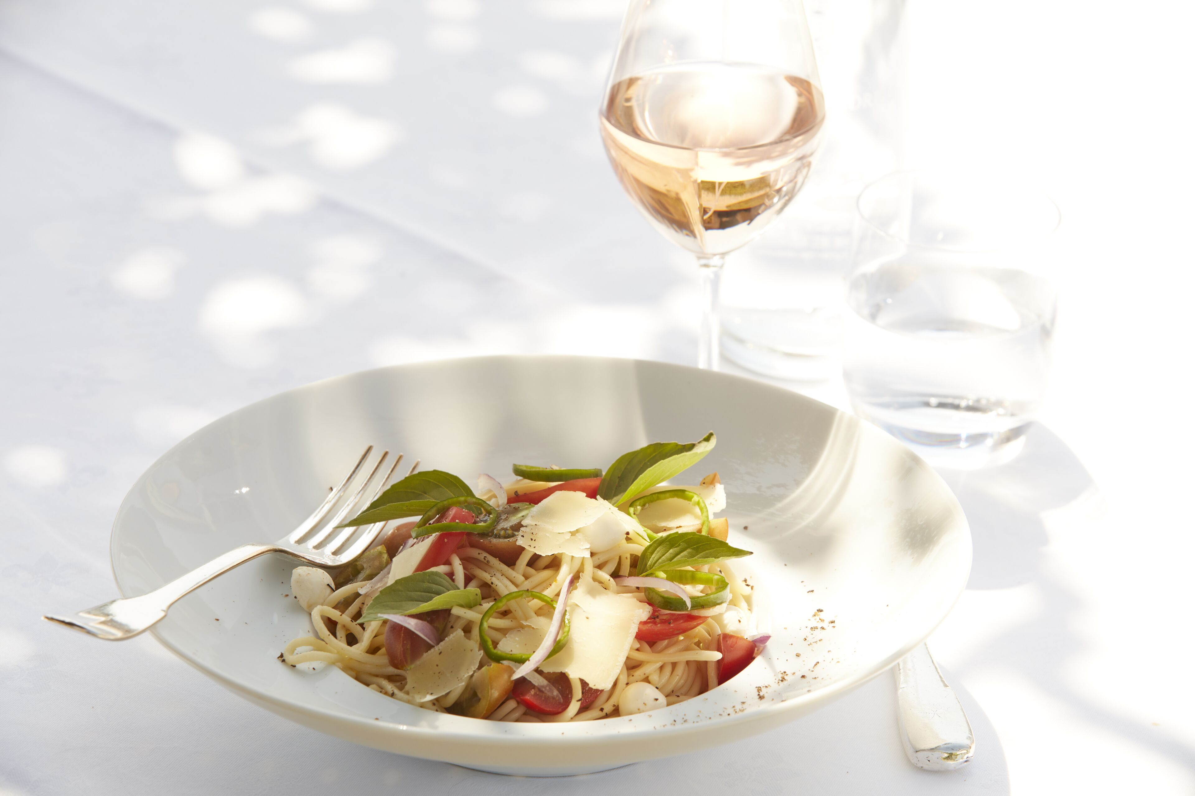 A white plate with a pasta dish, a fork, and a glass of wine on a table.