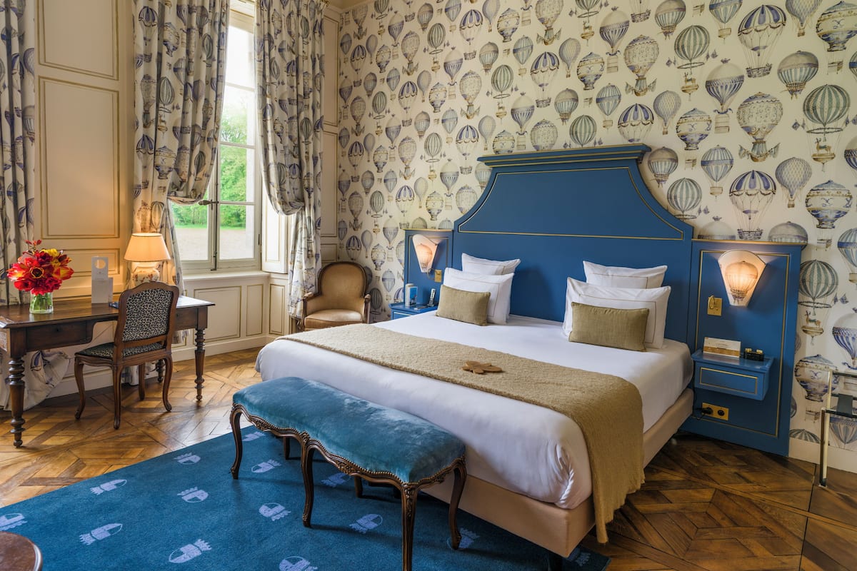 A bedroom with a large bed, a desk, and a chair. There is a window with curtains, a lamp, and a vase with flowers.