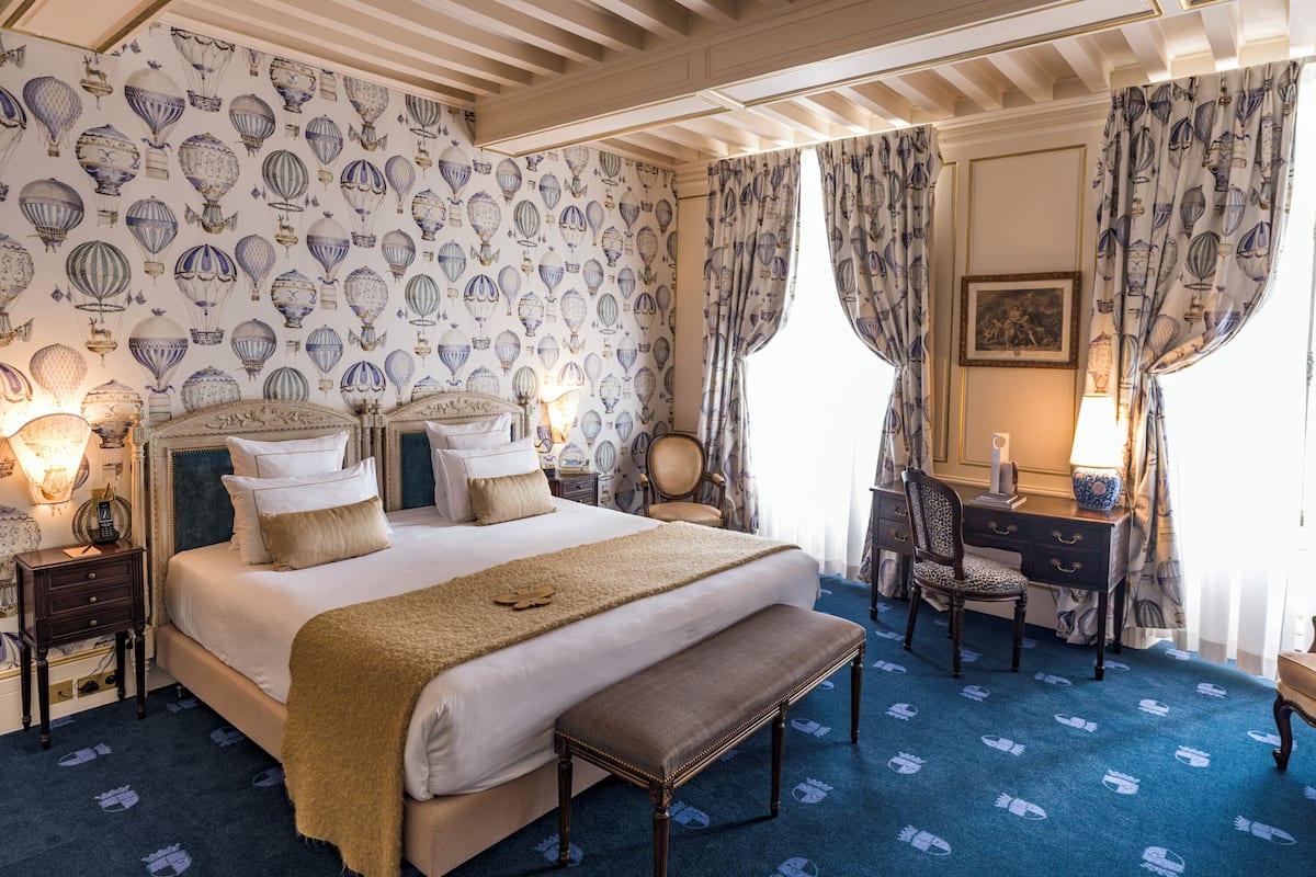 A hotel room with a large bed, a desk, and a chair. The room has a patterned wallpaper and blue carpet.