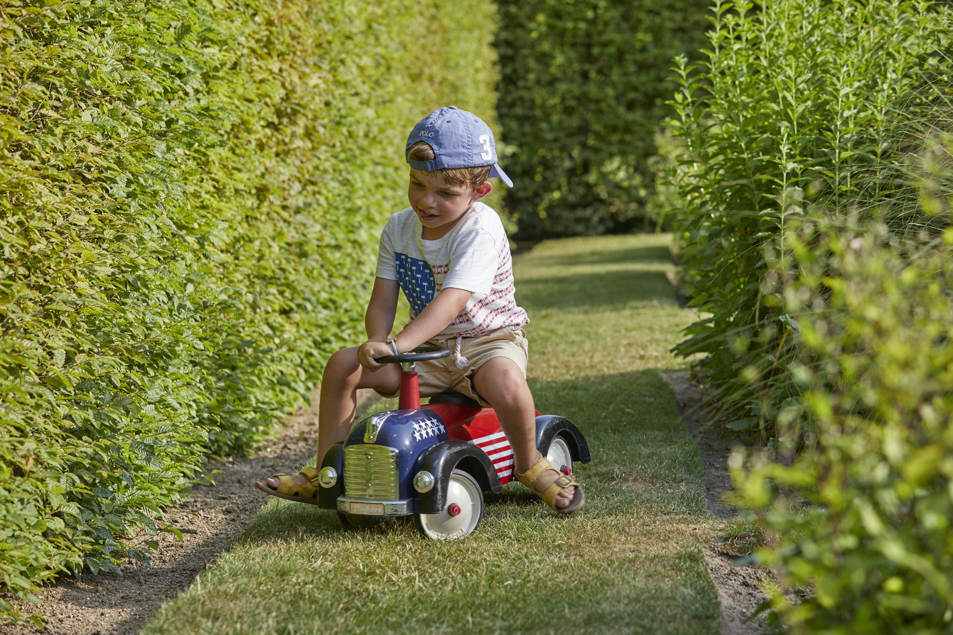 A child riding a toy car on a grassy path surrounded by hedges.