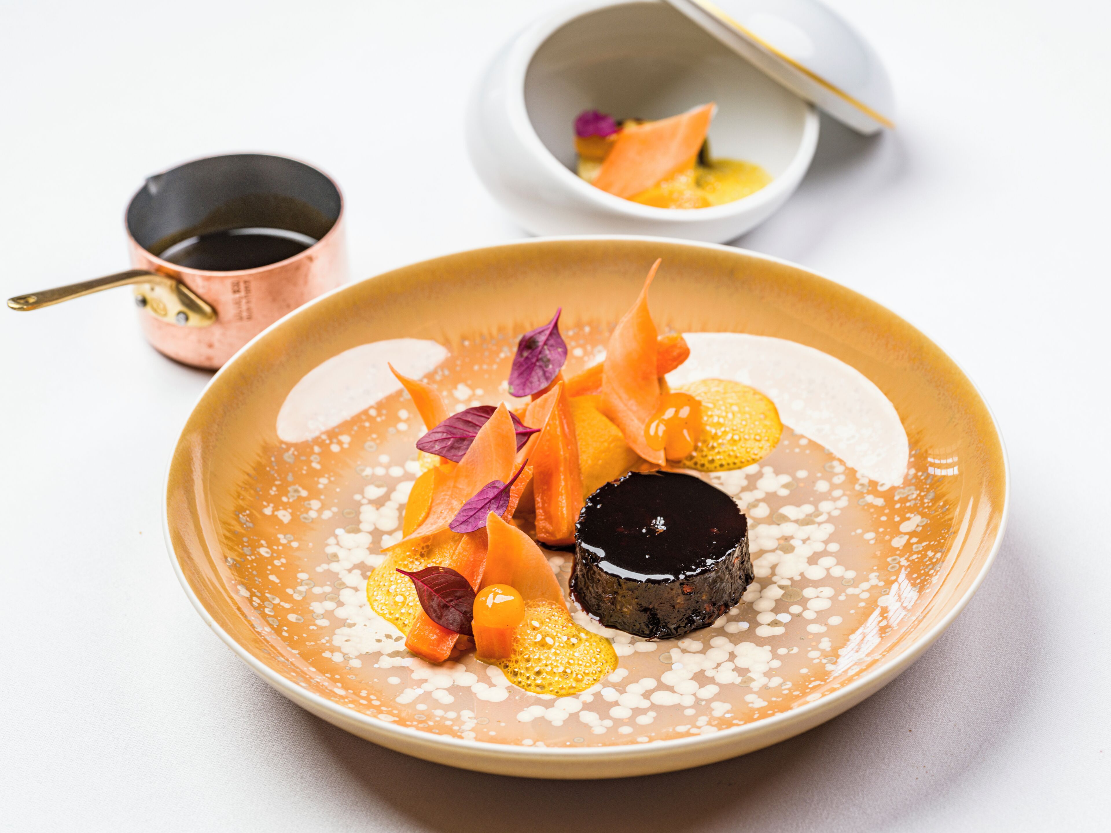 A dessert plate with a chocolate sphere, orange slices, and purple leaves on a beige plate.