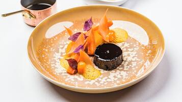 A dessert plate with a chocolate sphere, orange slices, and purple leaves on a beige plate.