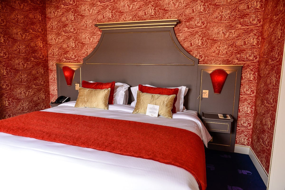 A hotel room with a large bed, red and gold pillows, a red throw blanket, and a decorative headboard.
