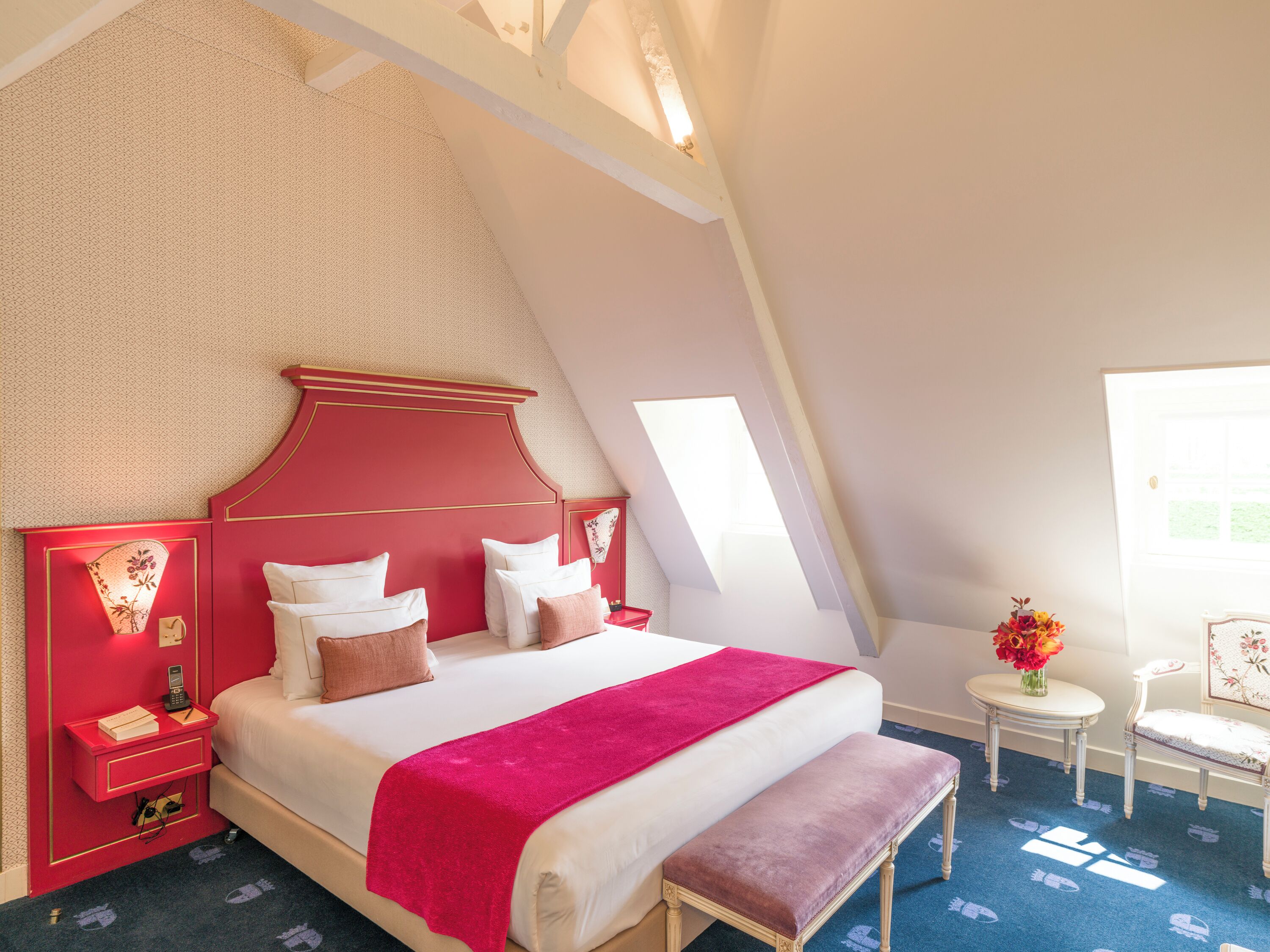 Un dormitorio con cabecera roja, una cama grande con ropa de cama blanca y rosa, un banco, una mesita con un jarrón de flores y una alfombra estampada.