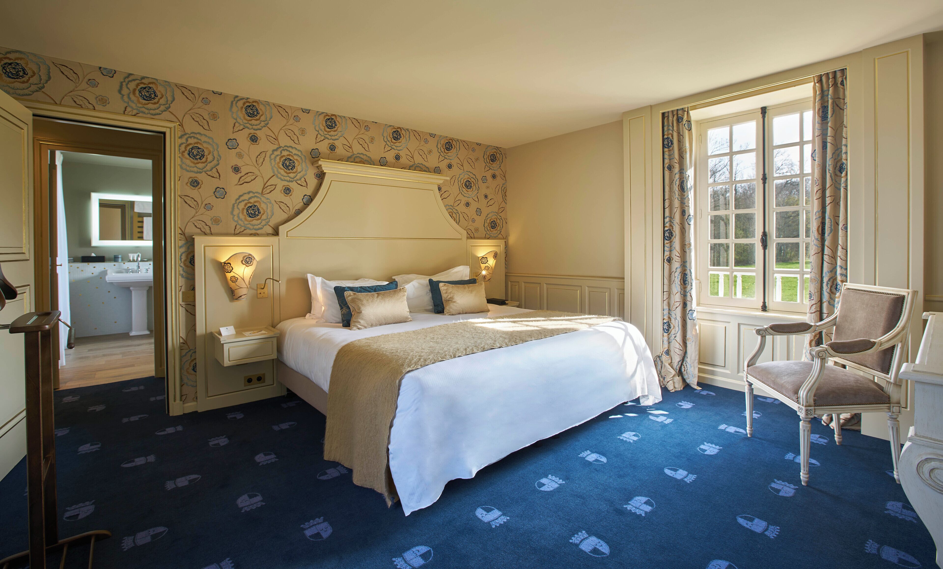 A hotel room with a large bed, a sitting chair, a nightstand, a mirror, and a window with curtains.