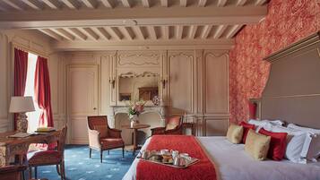 A hotel room with a large bed, a desk with a lamp, two chairs, a small table with a vase, and a tray with breakfast items.