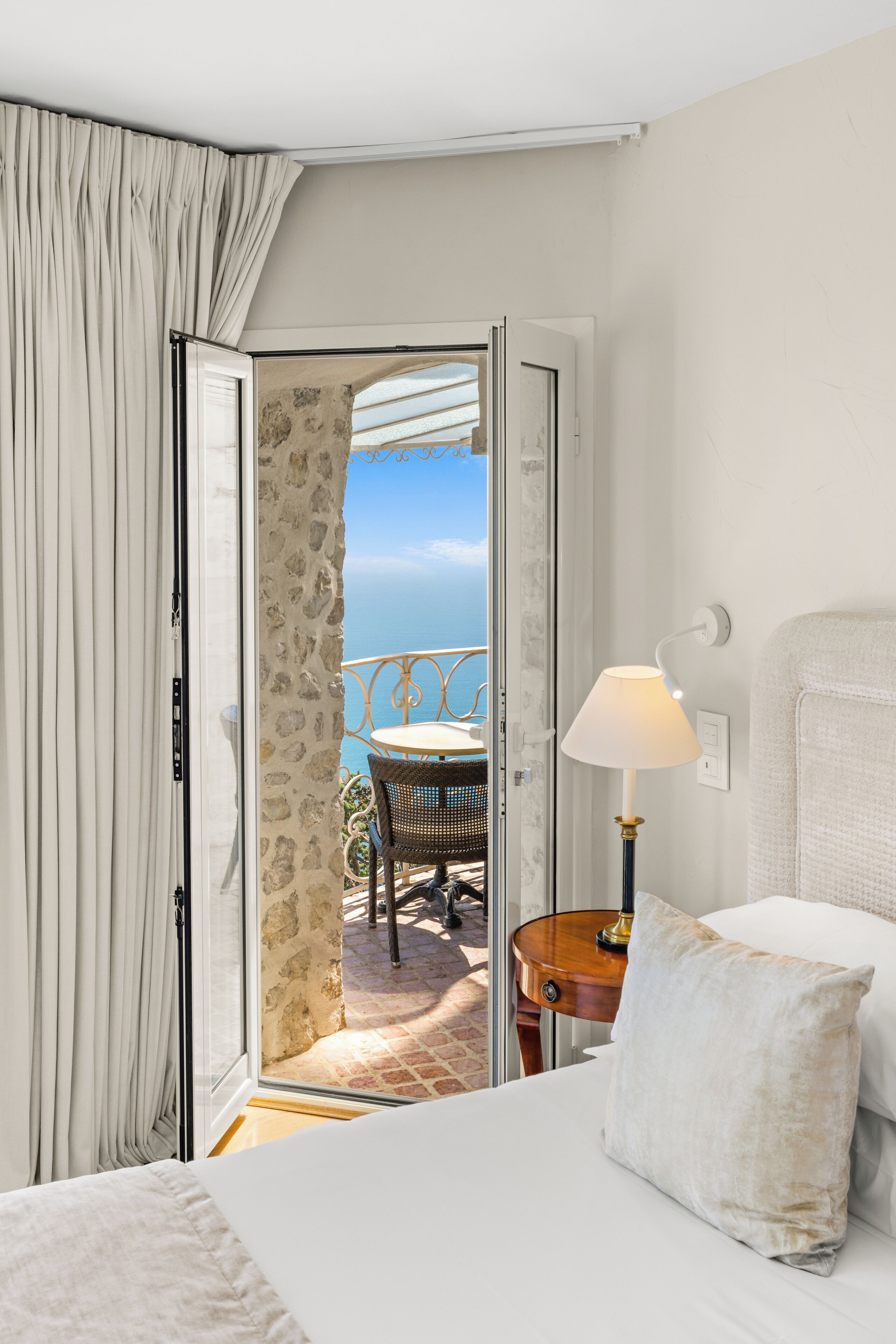 A hotel room with a bed, a nightstand, a lamp, a view of the sea through a glass door, and a balcony with a table and chairs.