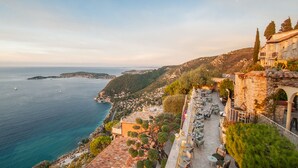 A coastal view with a terrace overlooking the sea, stone buildings, and a dining area with outdoor seating. - Château de La Chèvre d’Or (Èze)