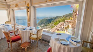 A dining area with tables and chairs, large windows with a view of a coastal landscape. - Château de La Chèvre d’Or (Èze)