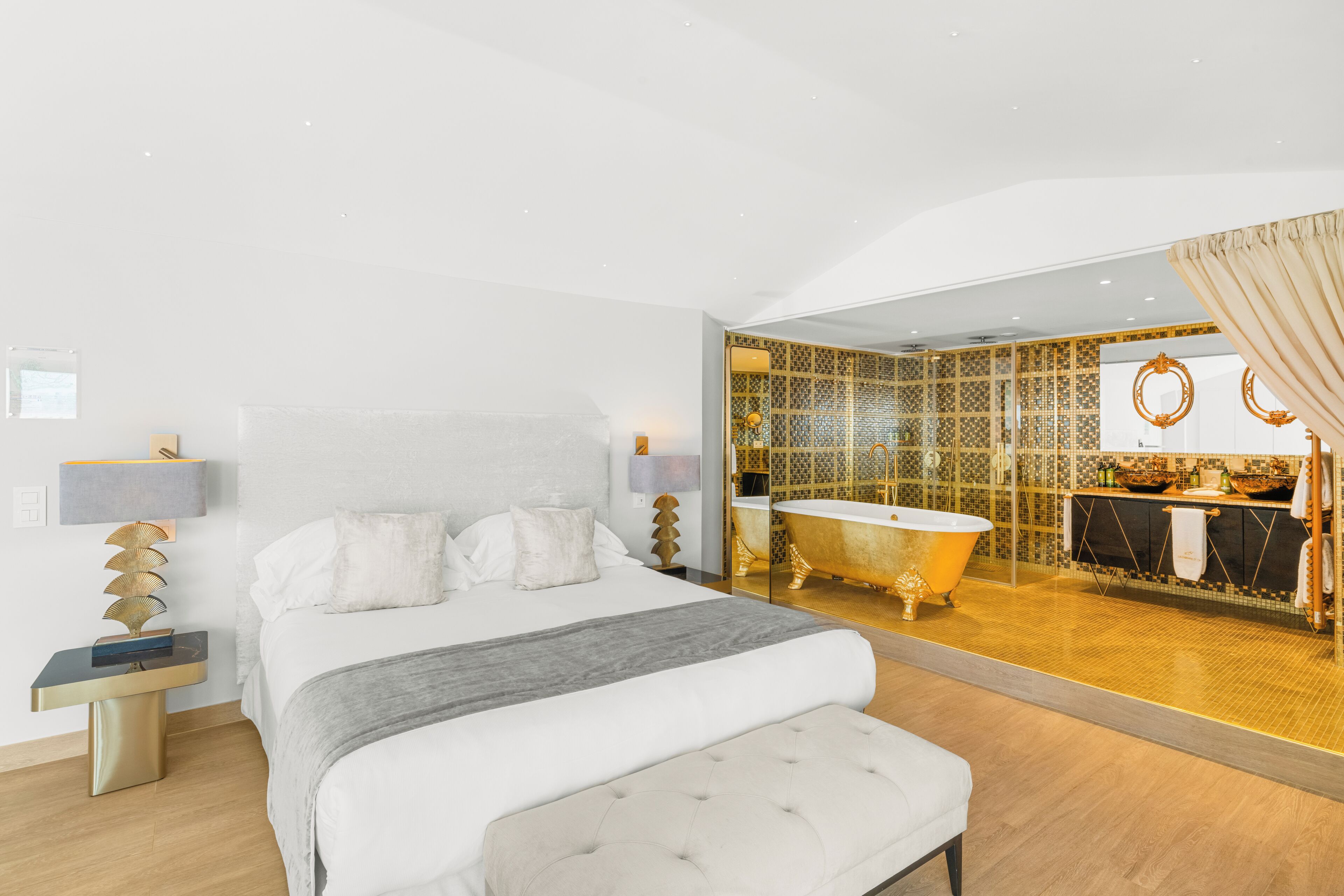 Een moderne slaapkamer met een groot bed, een nachtkastje, een lamp en uitzicht op een badkamer met een bad en een wastafel.