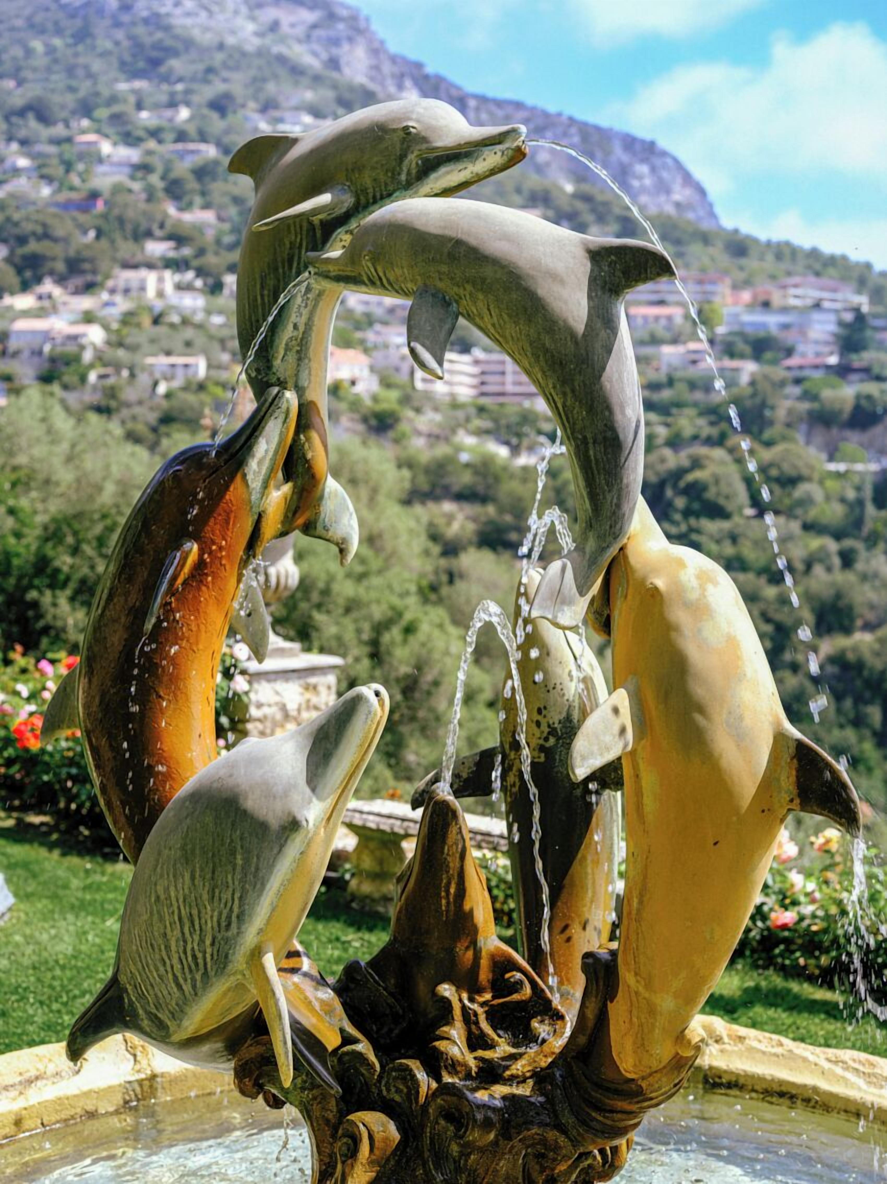 Une fontaine ornée de dauphins et de jets d’eau, dont le décor est constitué d’un paysage montagneux et d’une ville en arrière-plan.