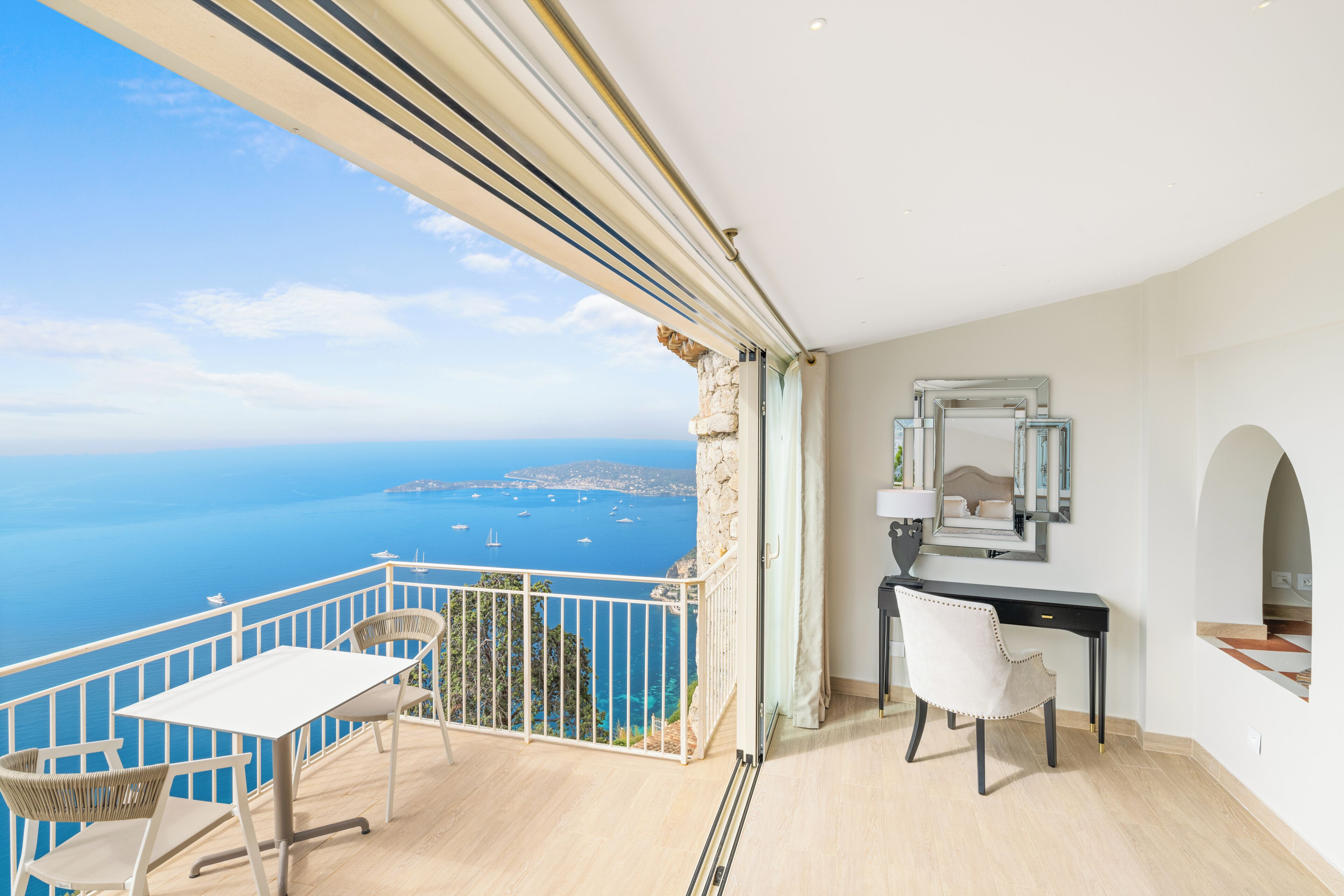 A balcony with a view of the sea and boats, a desk with a chair, and a stone wall.
