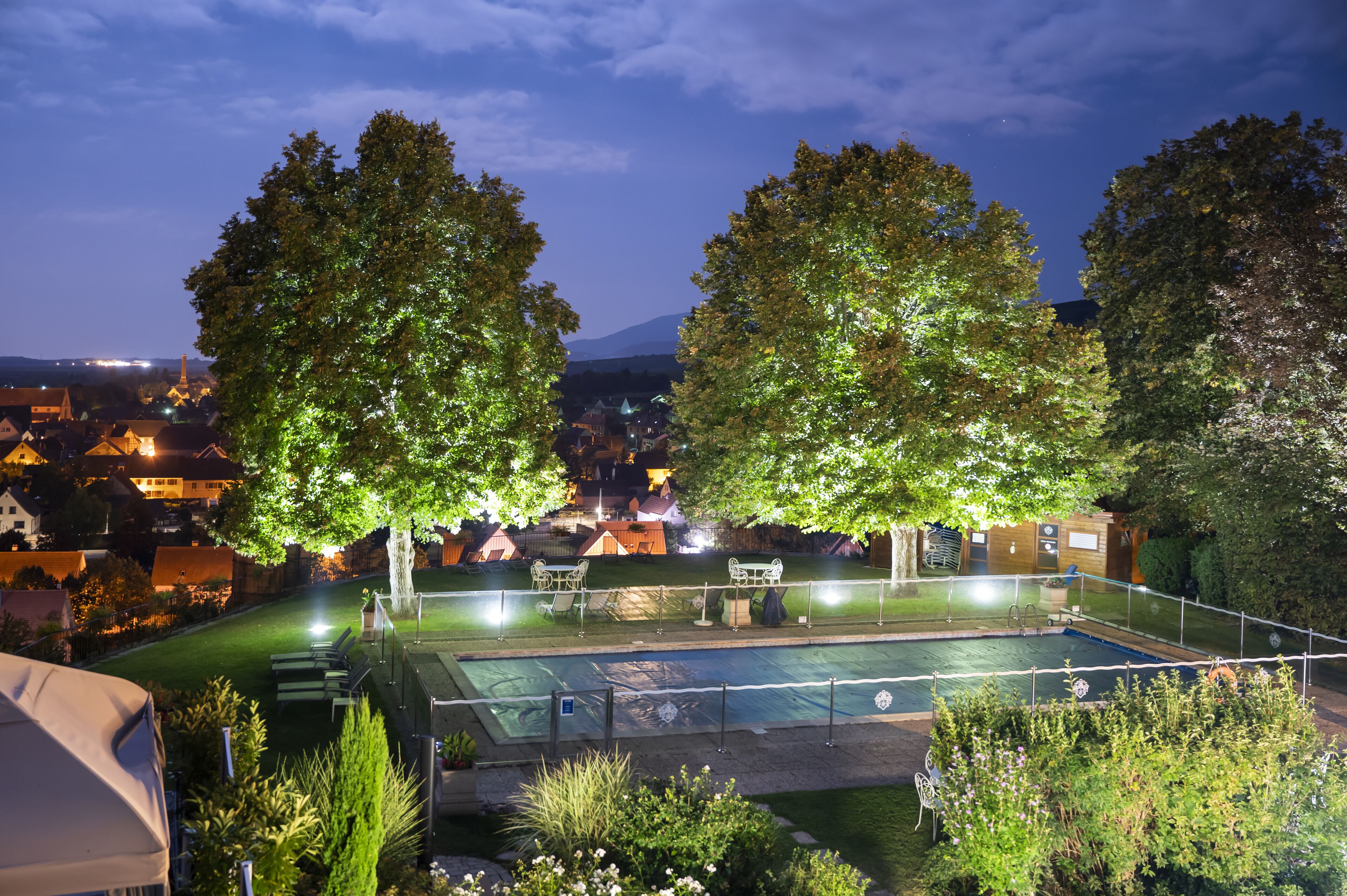 Piscina con valla, rodeada de árboles y vista a un pueblo de noche.