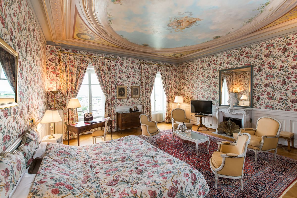 A hotel room with floral wallpaper, a large bed, two armchairs, a television, and a fireplace.
