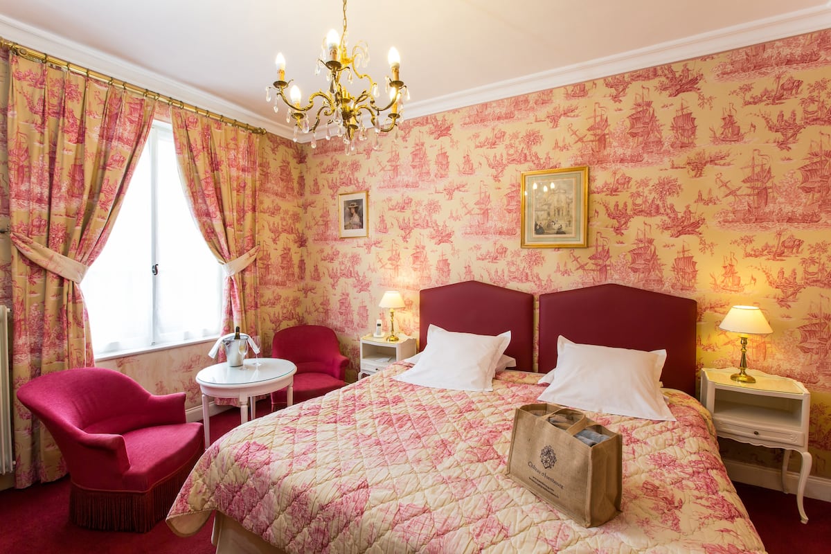 A hotel room with a large bed, two bedside tables, a red armchair, a small round table, and a chandelier.