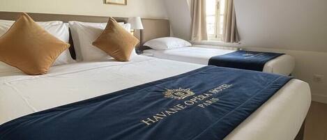 Ein Hotelzimmer mit zwei Betten, jedes mit einer blauen Tagesdecke und einem goldenen Wappen. Das Zimmer verfügt über Holzbalken an der Decke und ein Fenster mit Vorhängen.