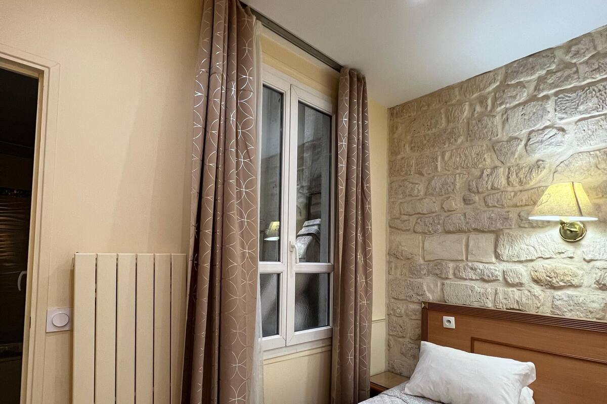 A bedroom with a bed, a window with curtains, a stone wall, and a wall-mounted lamp.