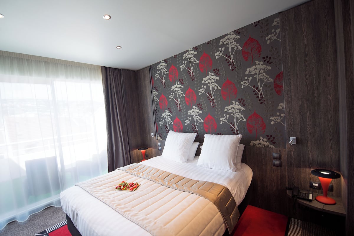 A hotel room with a large bed, a bedside table with a lamp, and a window with sheer curtains.