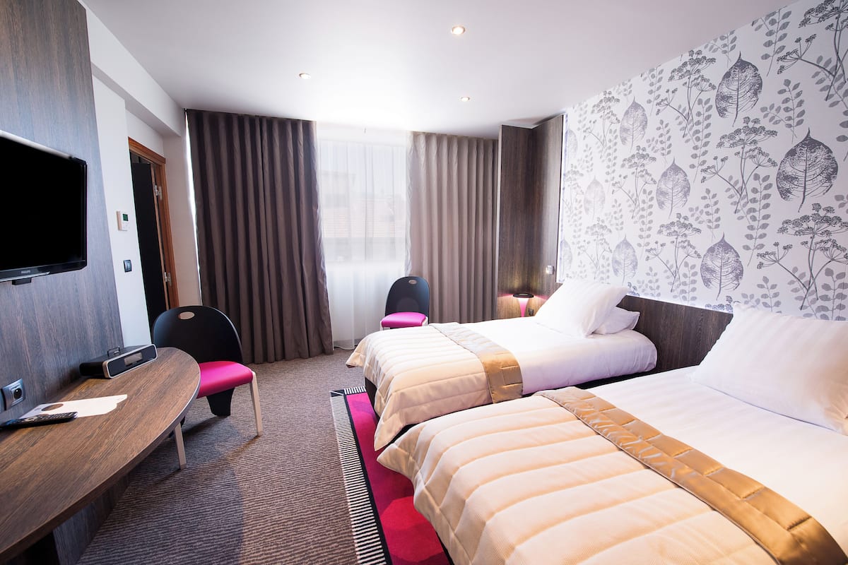 A hotel room with two beds, a desk with a chair, a TV, and a large window with curtains.