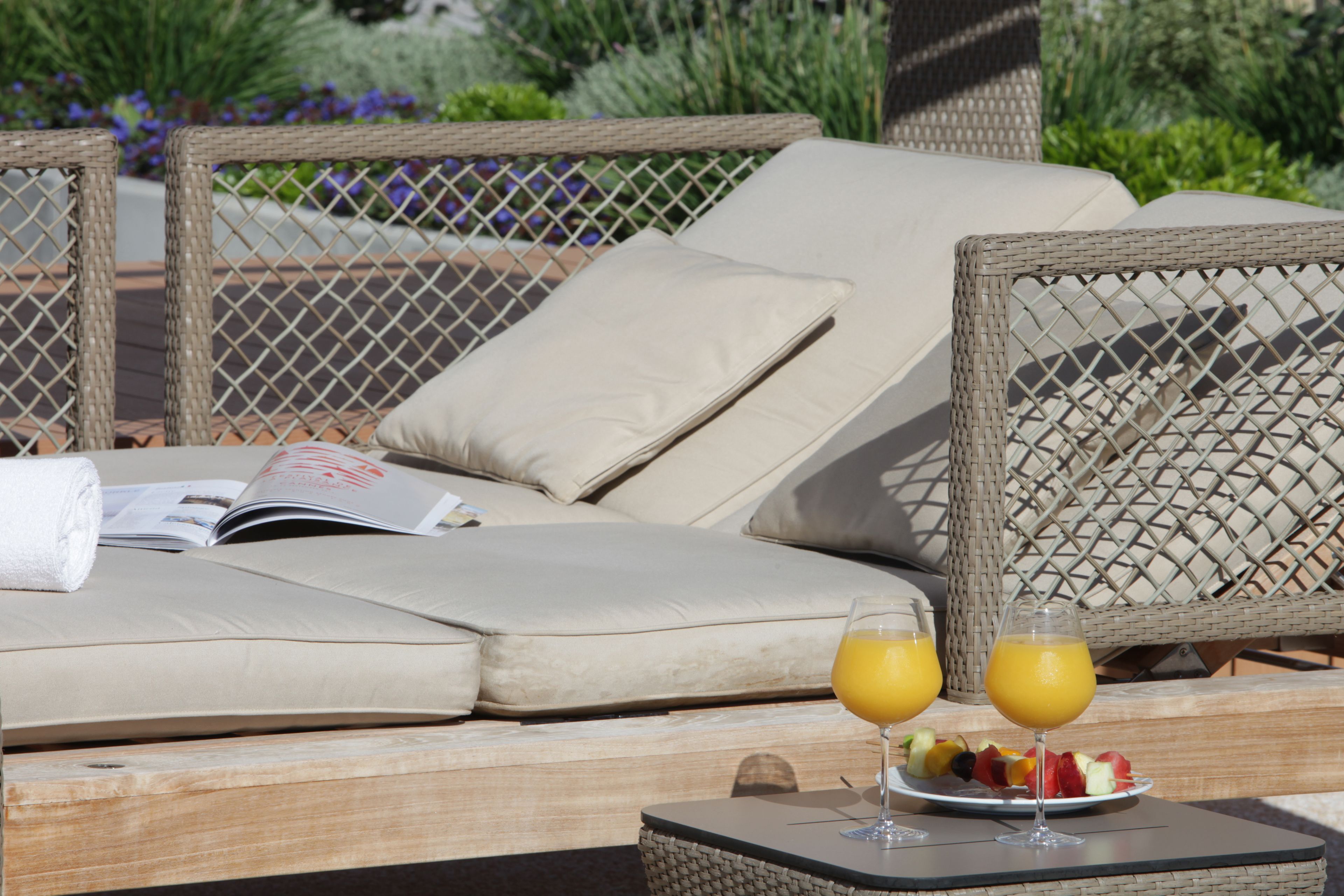 A wicker lounge chair with a cushion, a magazine, and a tray with fruit and drinks.