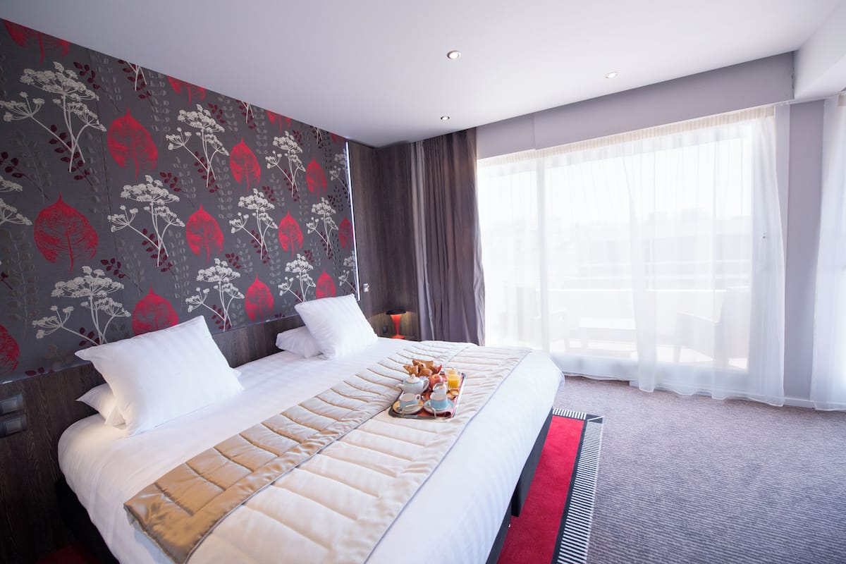 A hotel room with a large bed, a red carpet, and a window with sheer curtains.