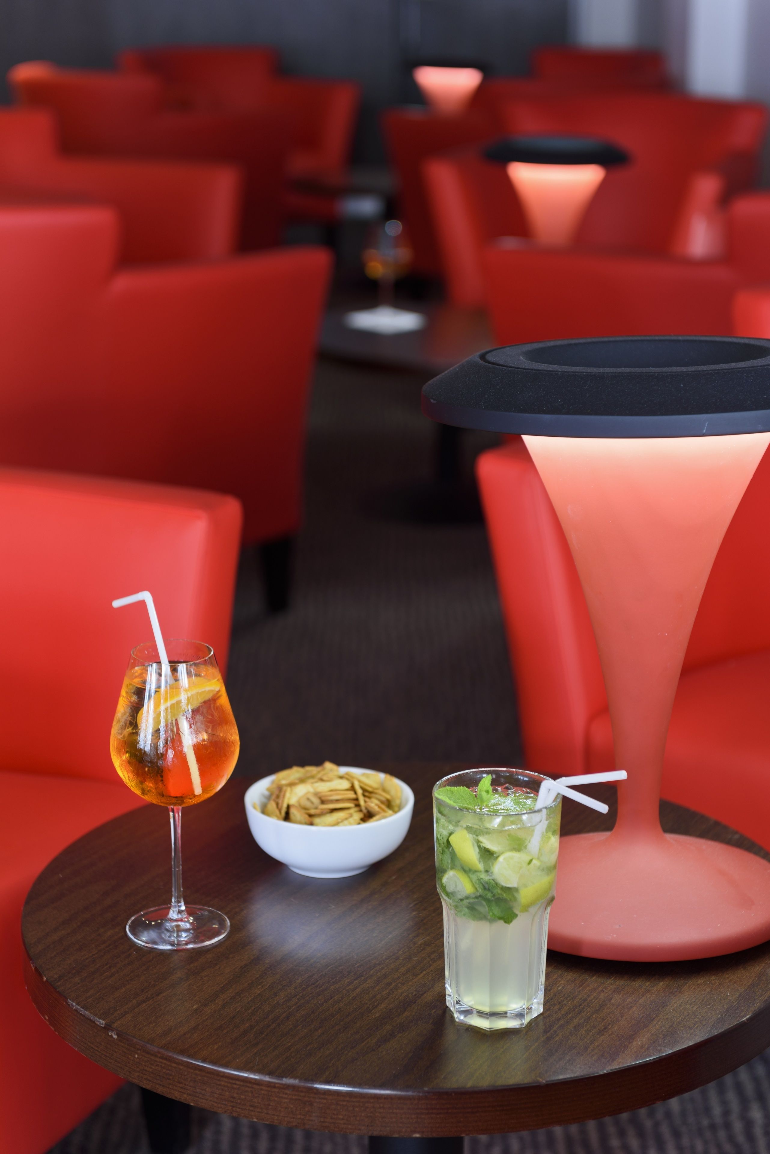 Ein Tisch mit einem Cocktail, einem Wasserglas mit Trinkhalm und einer Schale mit Snacks in einem modernen Ambiente mit roten Stühlen und einer außergewöhnlichen Tischlampe.