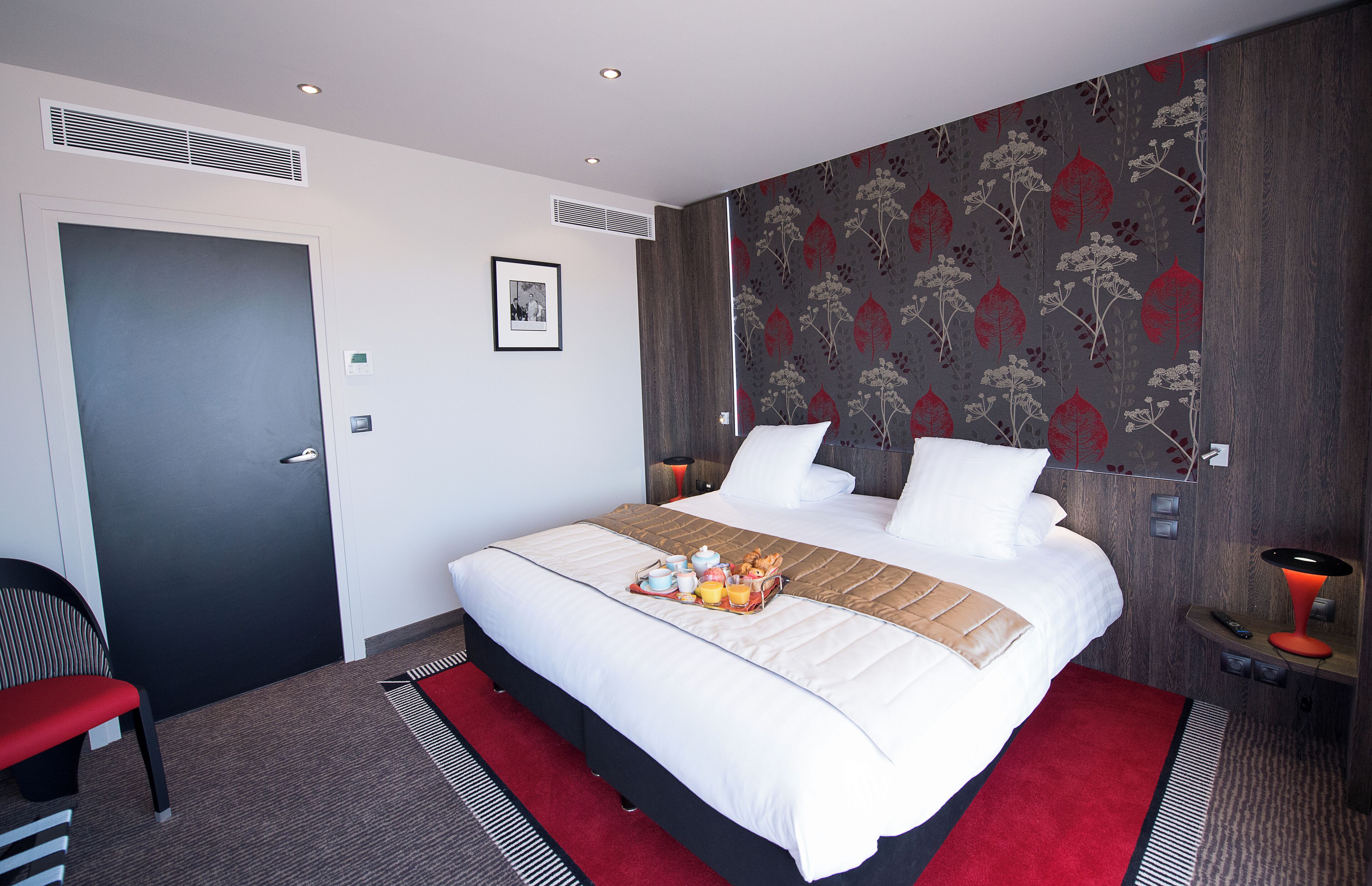 A modern hotel room with a large bed, a red and black striped rug, a small table with a lamp, and a framed picture on the wall.