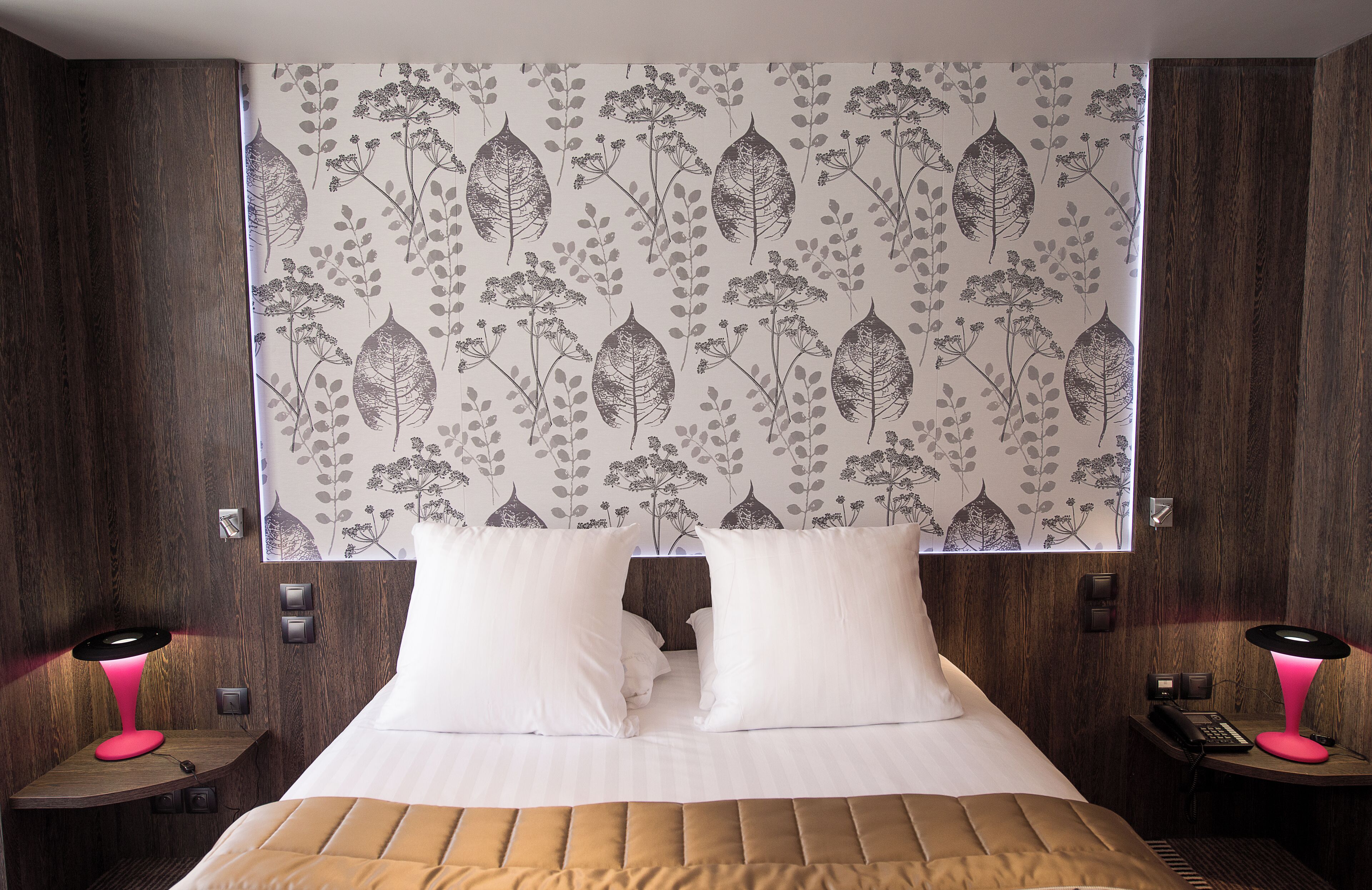 Ein Hotelzimmer mit einem großen Bett, zwei Nachttischen, einem Schreibtisch, einem Fenster mit einem vorfensterartigen Vorhang mit Blattmuster und einem an der Wand montierten Telefon.