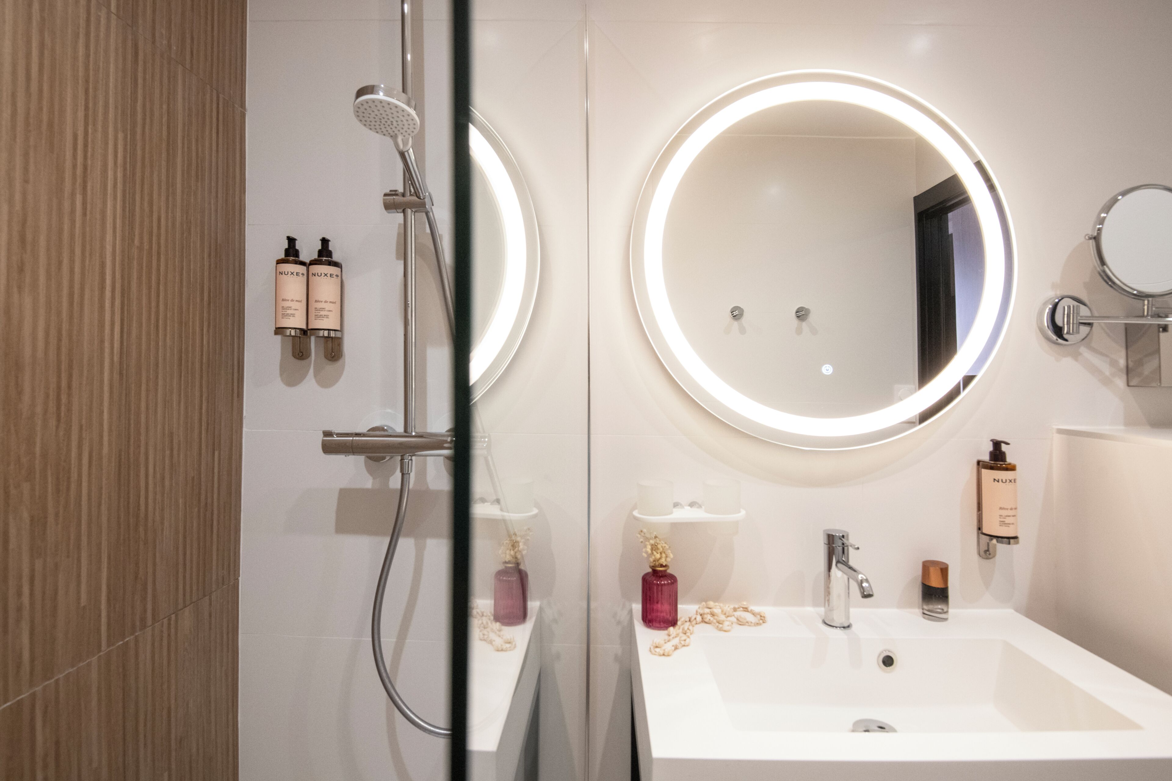 A modern bathroom with a glass-enclosed shower, a round mirror with integrated lighting, and a white sink with toiletries.
