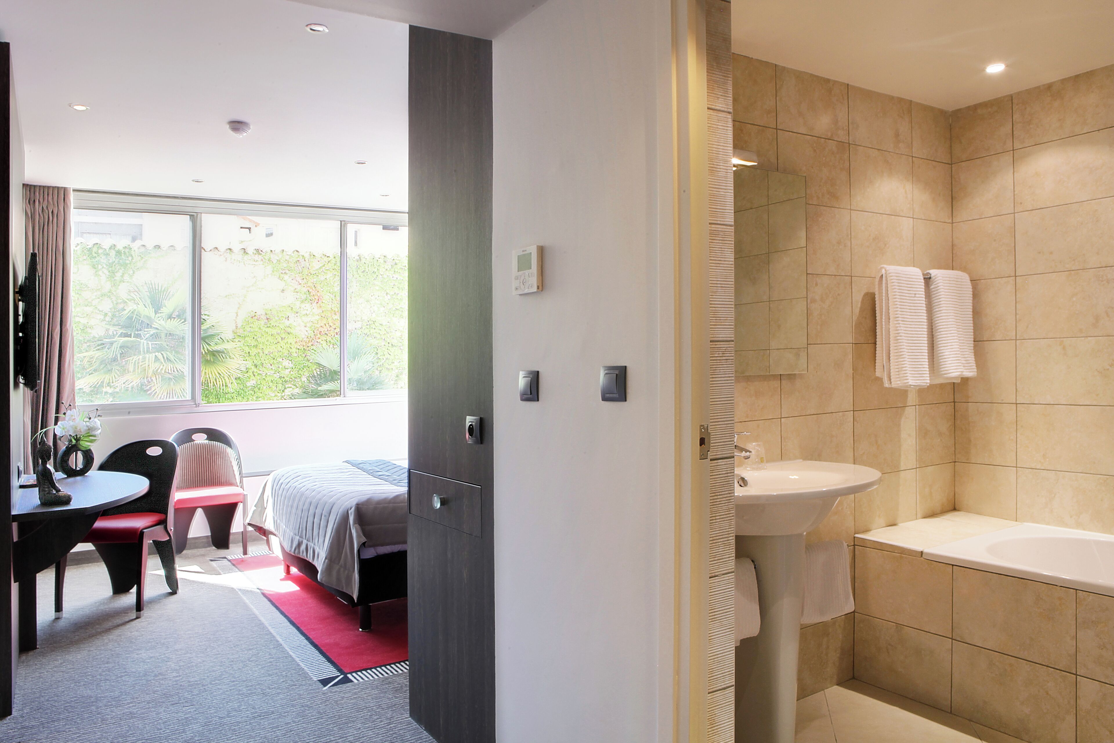 A modern hotel room with a bed, a desk with a chair, a bathroom with a sink and bathtub, and a window with a view of greenery.