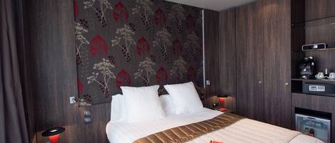 A hotel room with a large bed, a bedside table with a lamp, a small refrigerator, and a shelf with a coffee maker.