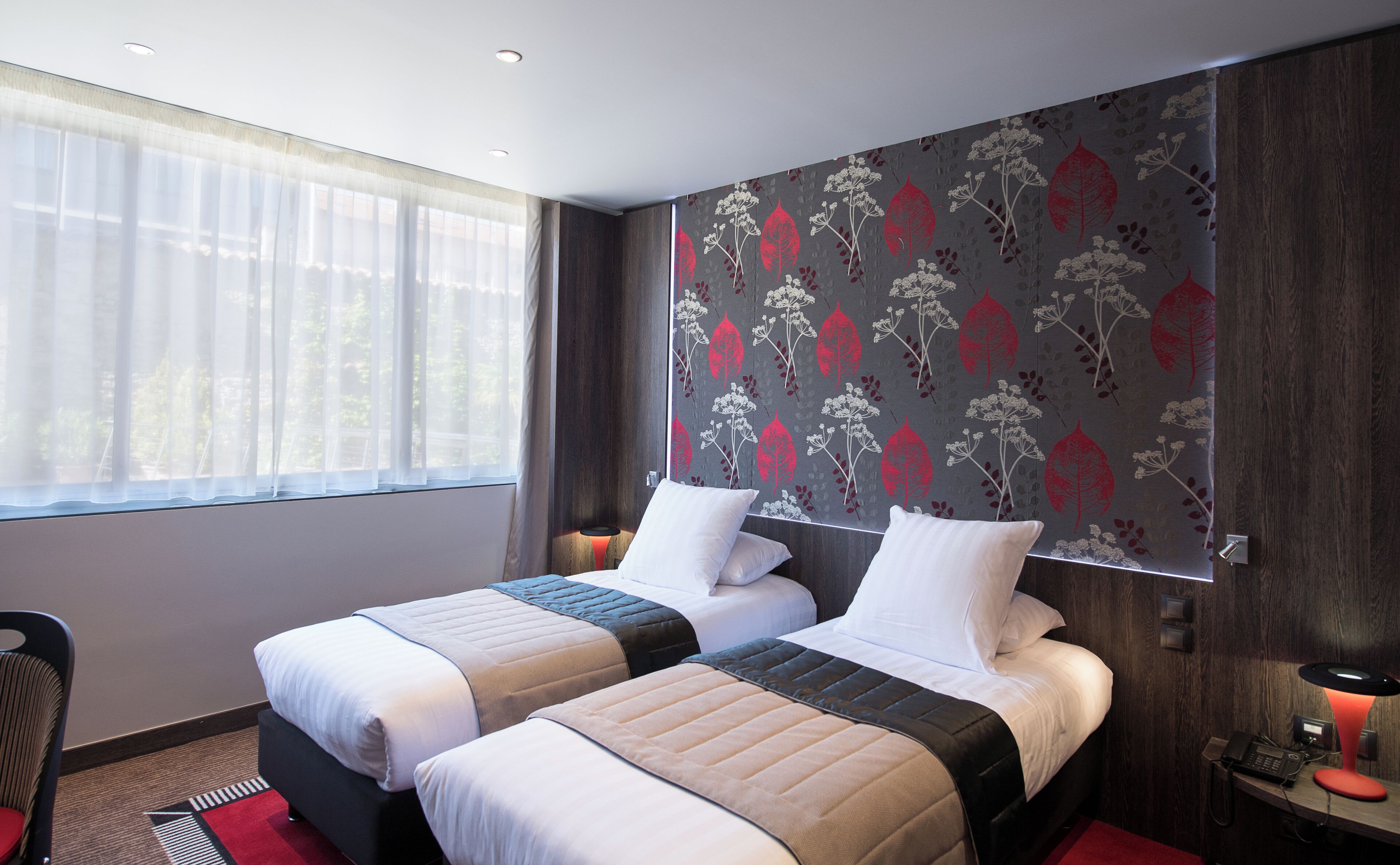 A hotel room with two beds, a window with sheer curtains, a wall with a floral wallpaper, and a desk with a phone and lamp.