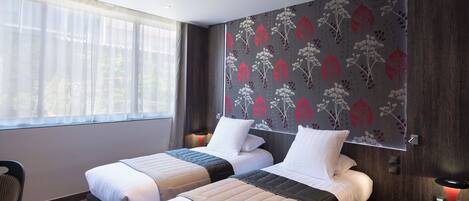 A hotel room with two beds, a window with sheer curtains, a wall with a floral wallpaper, and a desk with a phone and lamp.