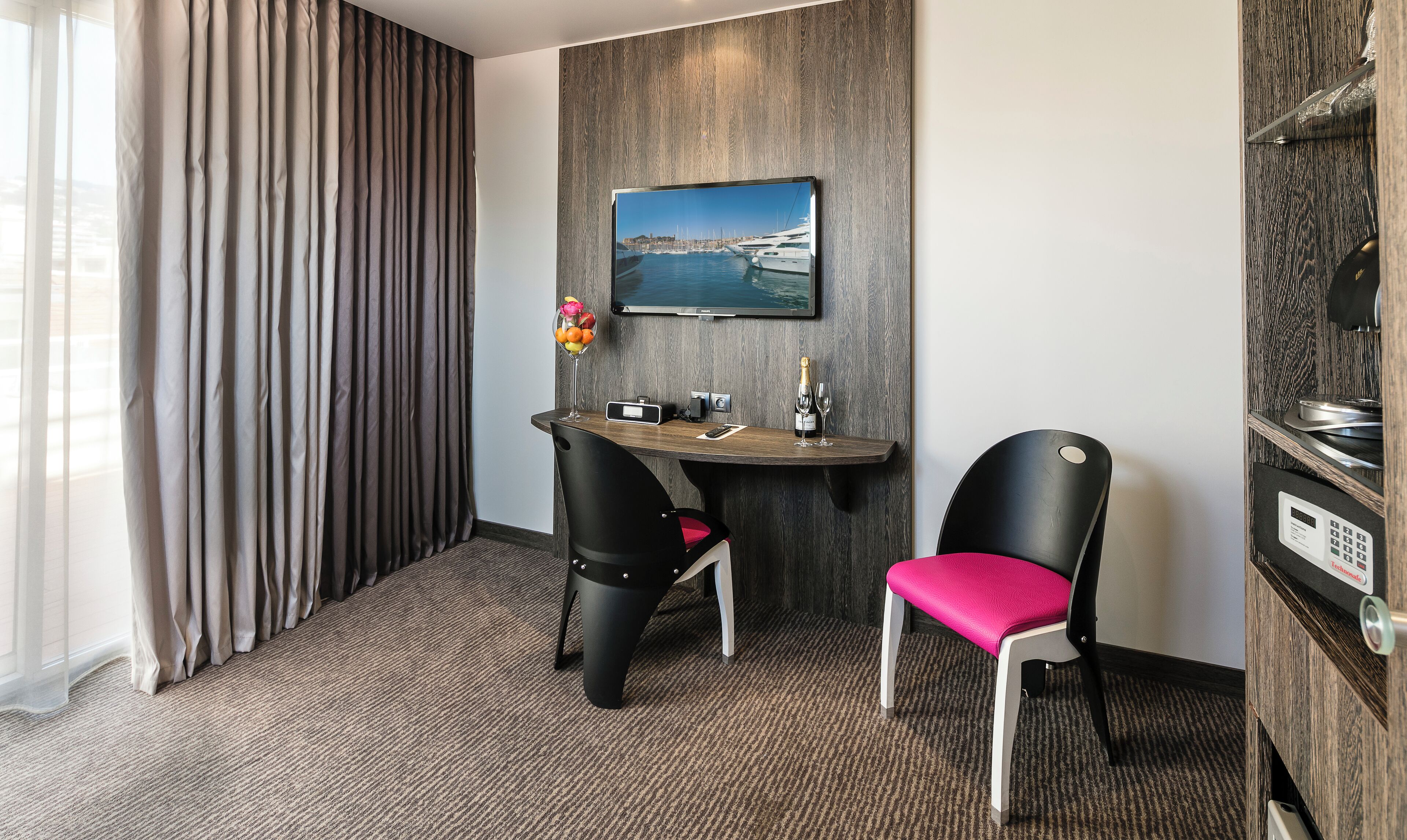 A hotel room with a dining table, two chairs, a painting of a marina, a minibar, and a large window with curtains.