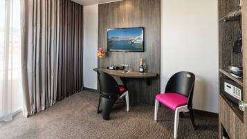 Habitación de hotel con una mesa de comedor, dos sillas, un cuadro de un puerto deportivo, un minibar y un ventanal con cortinas.
