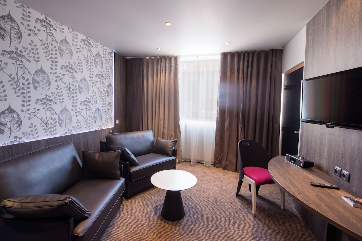 A modern hotel room with a sofa, a small round table, a TV, and a window with curtains.