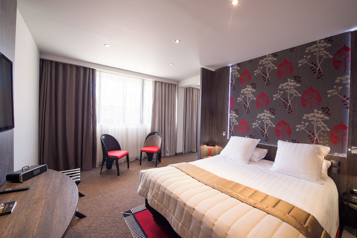 A hotel room with a large bed, two chairs, a TV, and a desk with a remote control.