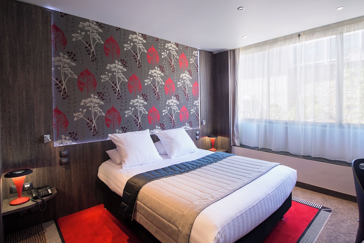A modern hotel room with a large bed, a desk with a lamp, a chair, and a window with sheer curtains.