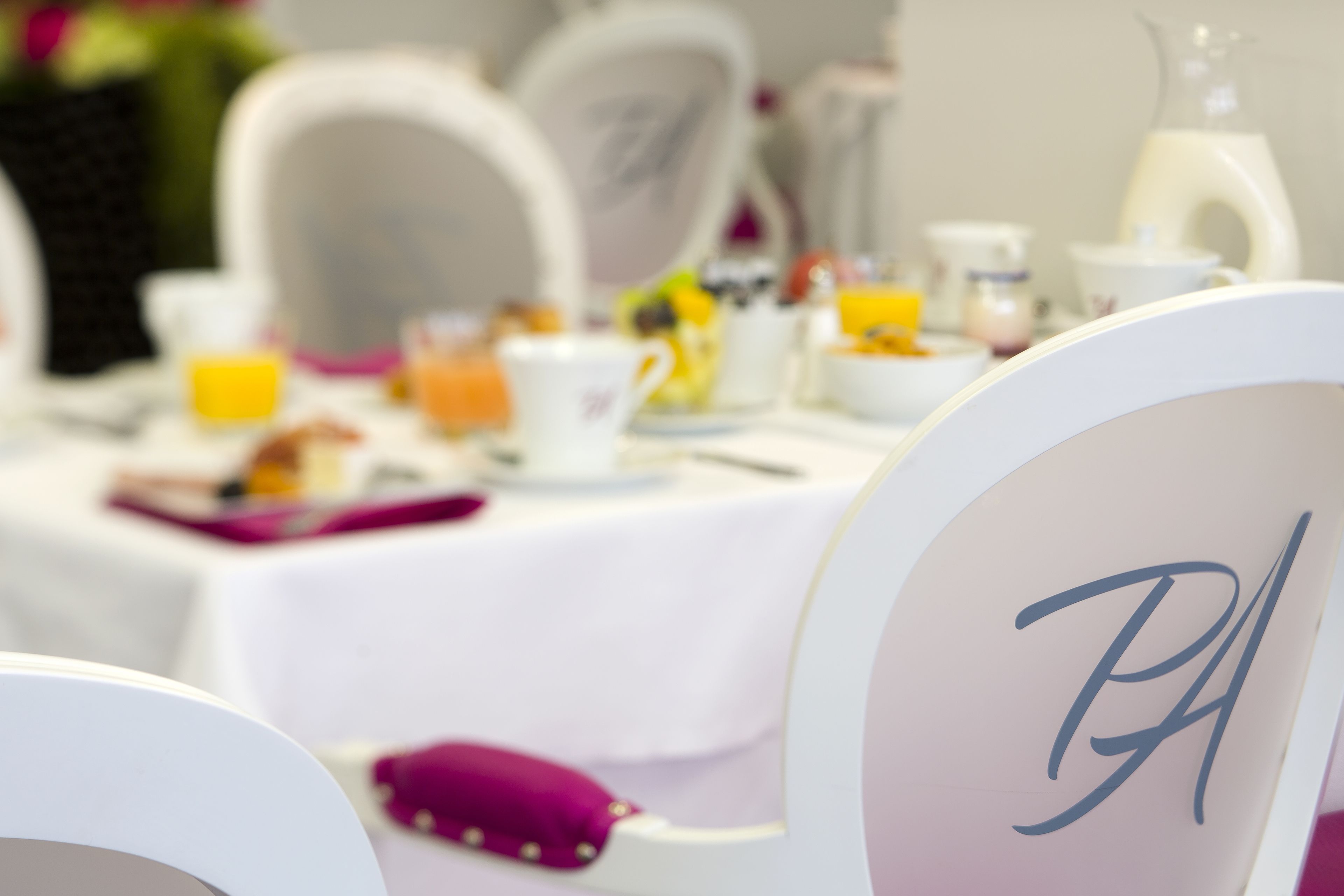 A dining table with a white tablecloth, chairs with monogrammed headrests, and various breakfast items including a glass of orange juice.