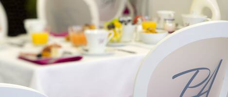 A dining table with a white tablecloth, chairs with monogrammed headrests, and various breakfast items including a glass of orange juice.