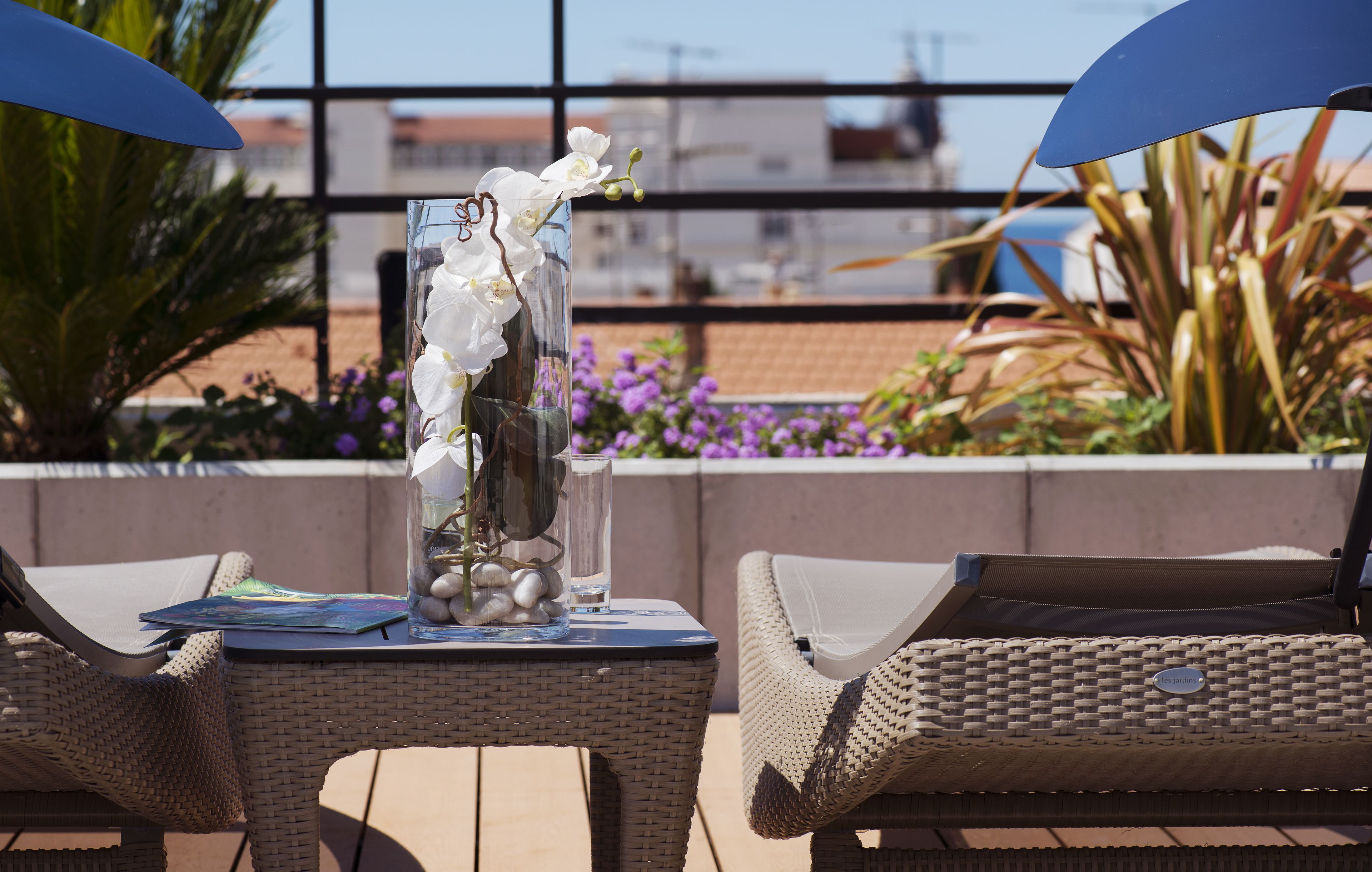A balcony with wicker chairs, a table with a vase of white flowers, and a view of the sea and buildings.