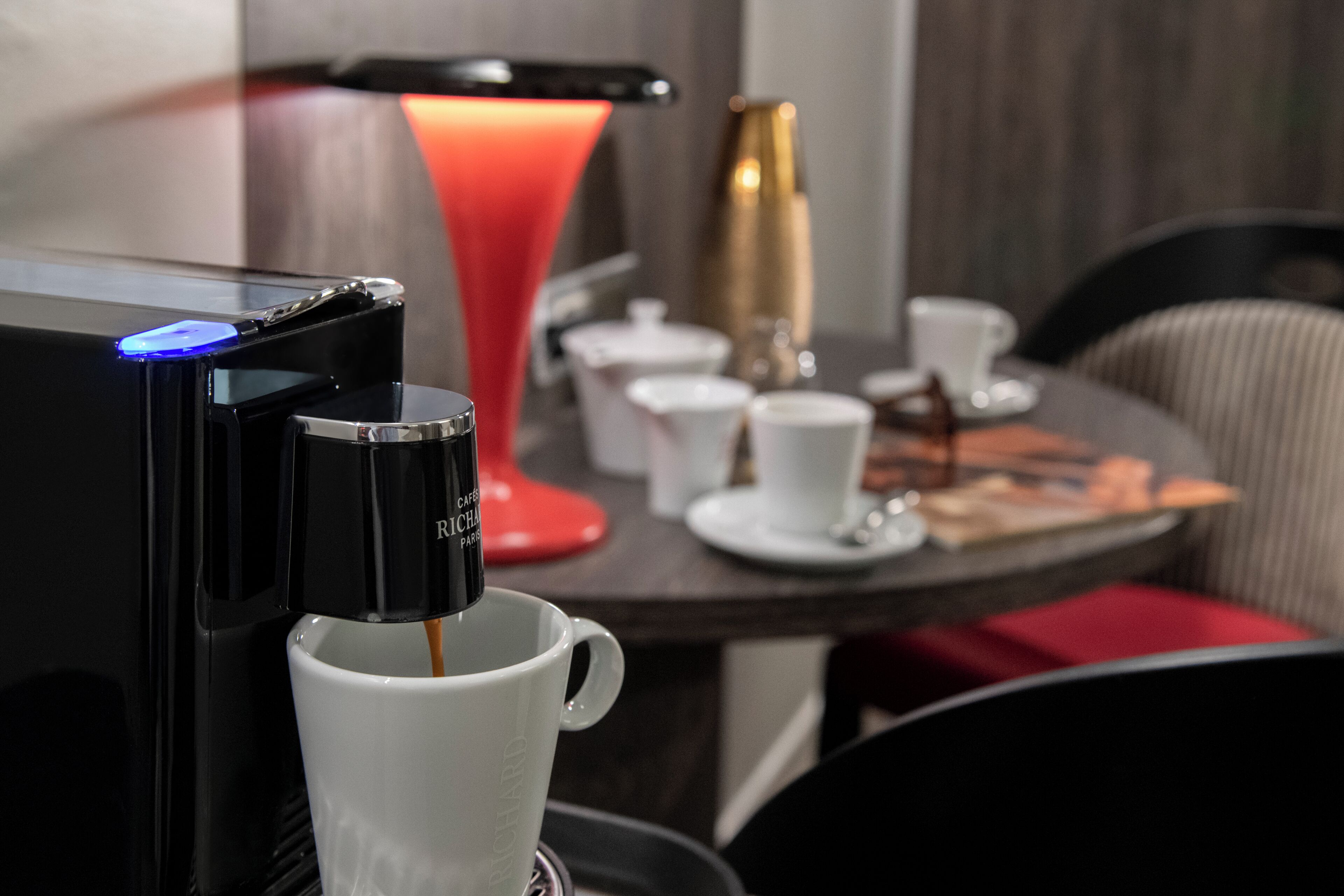 En kaffemaskine serverer kaffe i et hvidt krus, en rød lampe og et lille bord med krus og en dekorativ flaske.