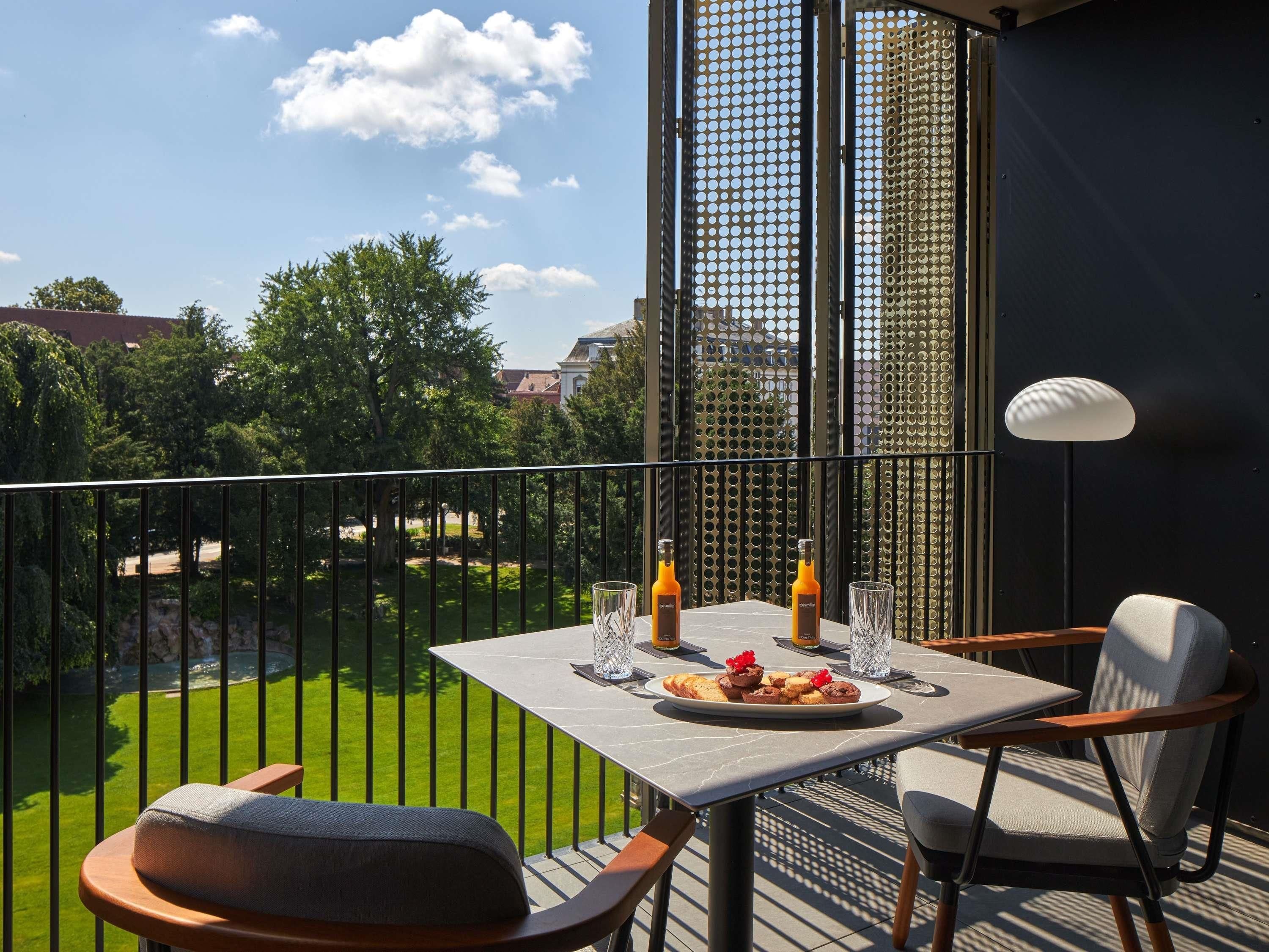 Een balkon met een tafel gedekt voor twee personen, twee flesjes sinaasappelsap, een bord gebak en een lamp.