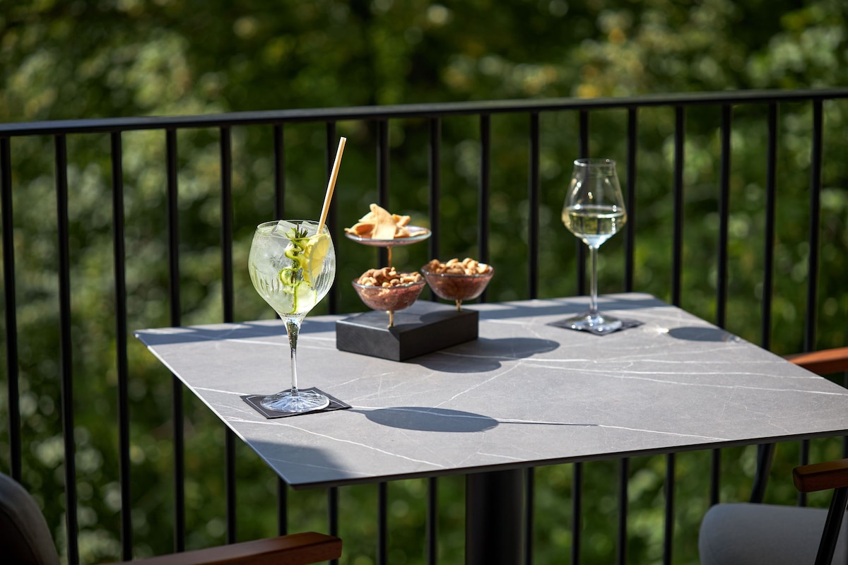 Une table sur une terrasse, agrémentée d’une barrière en métal, sur laquelle on trouve un verre à martini, un verre à vin et un bol de noix.