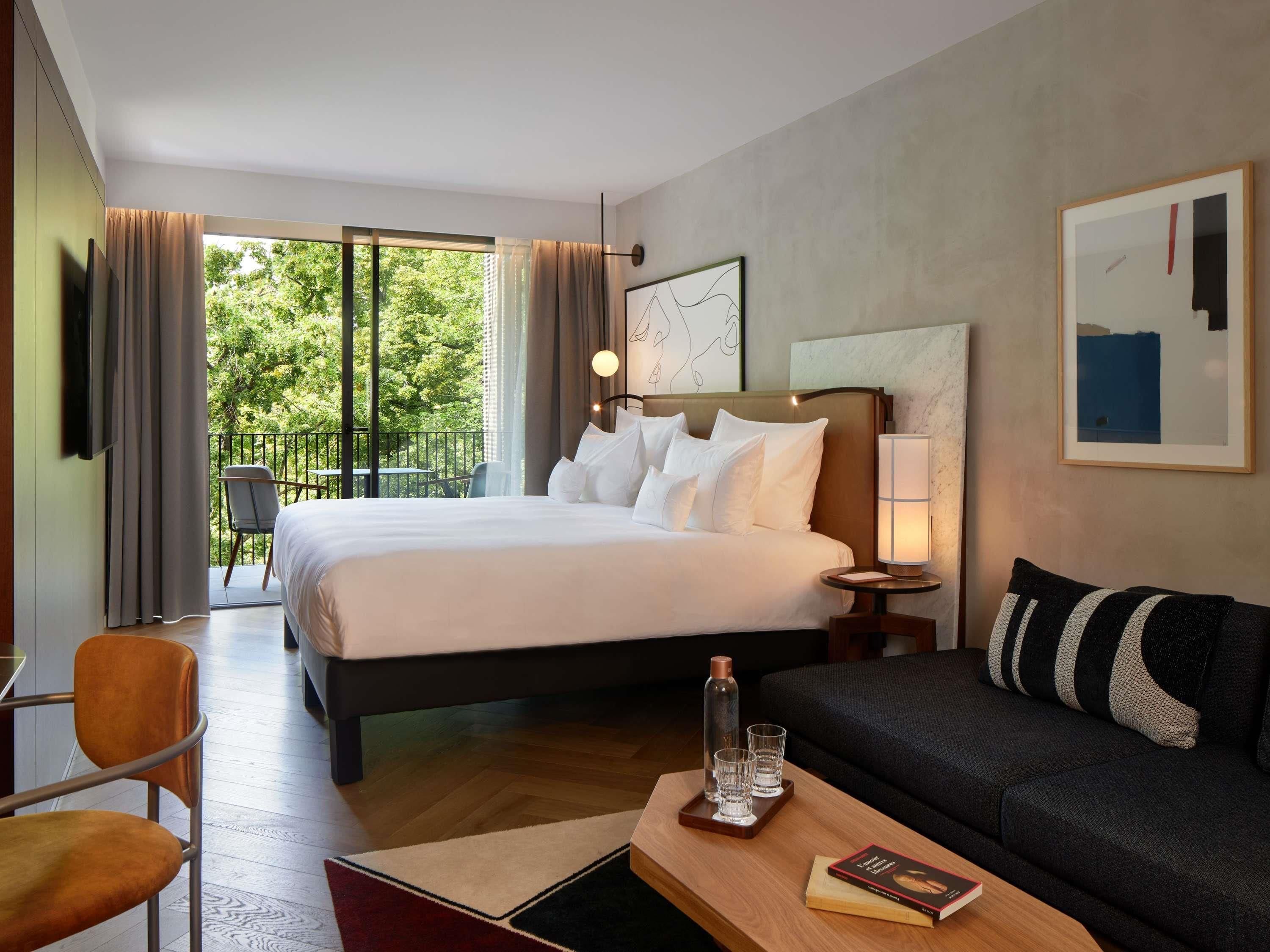 A hotel room with a bed, a sofa, a table with glasses and a book, a lamp, and a painting on the wall.
