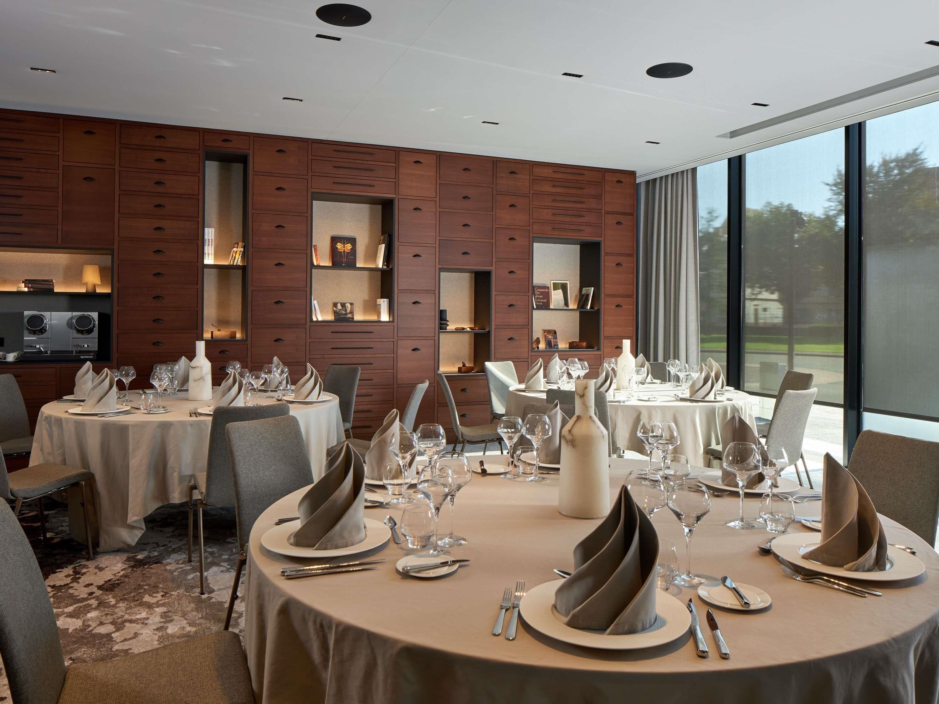 Un comedor con mesas redondas, elegantemente dispuestas con platos, vasos y cubiertos. Hay estanterías con libros y objetos decorativos. La habitación tiene un diseño moderno con paneles de madera y ventanas amplias.