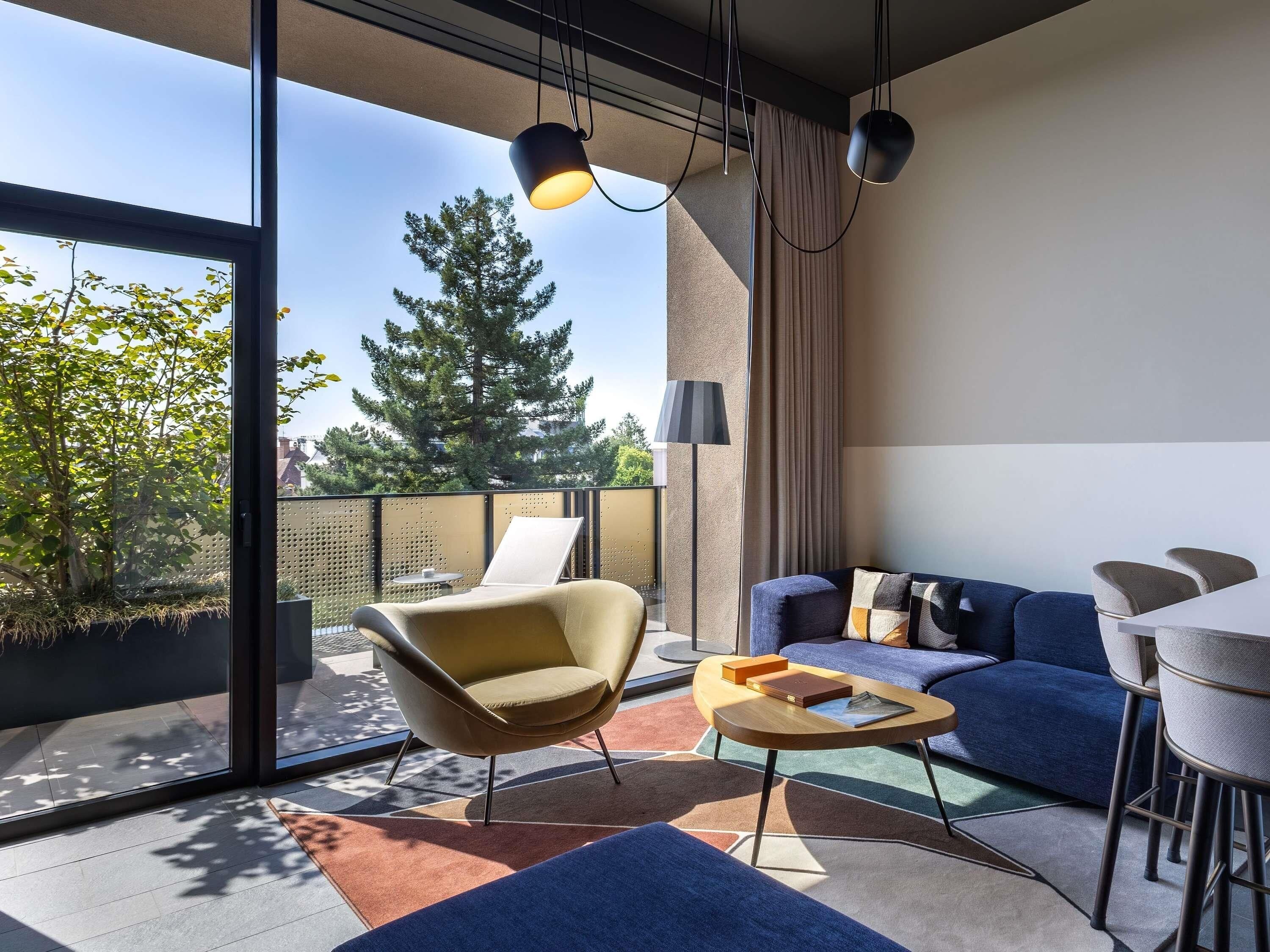 A modern living room with a sofa, armchair, and coffee table, large windows with a view of trees, and a balcony.