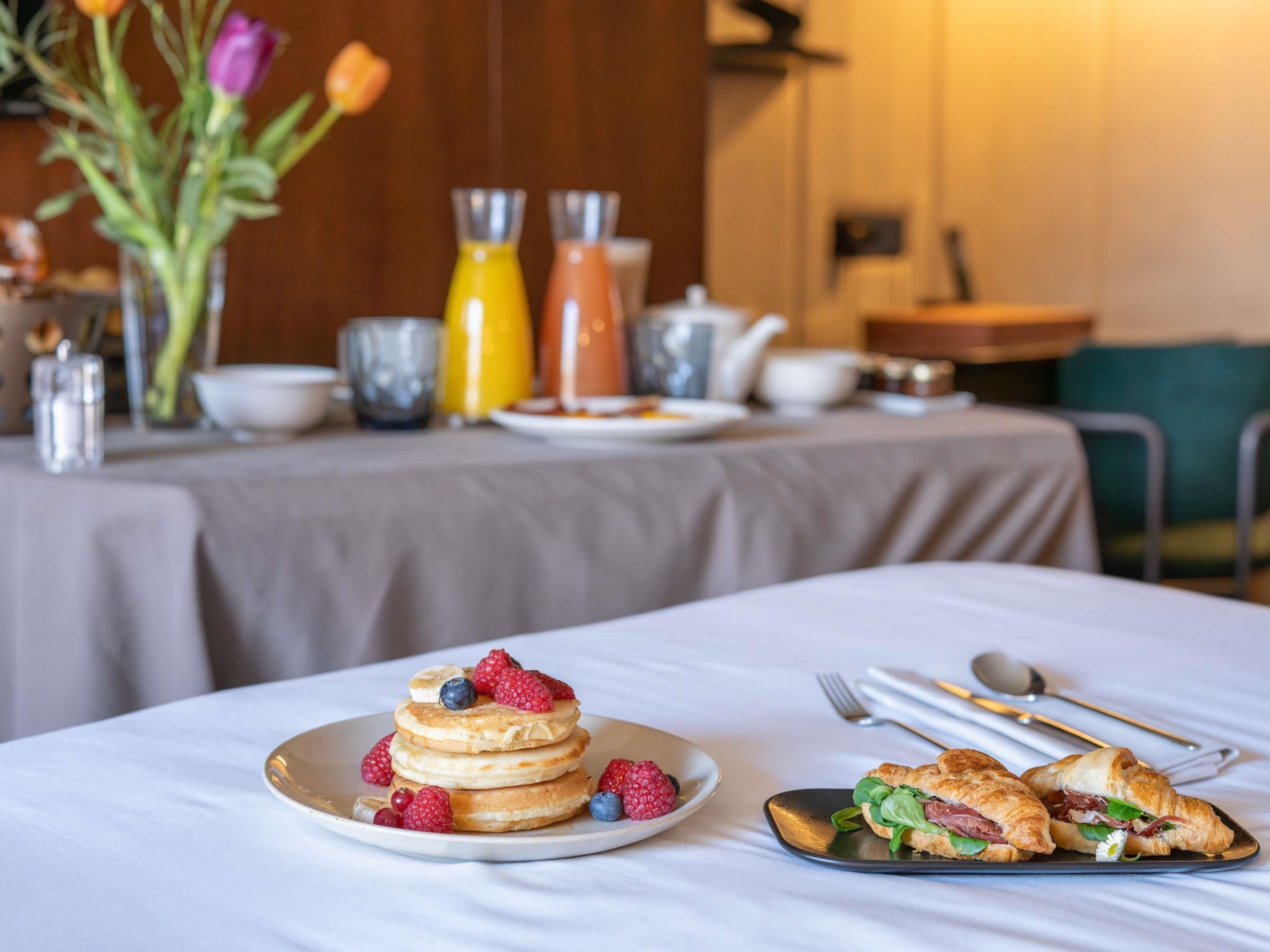 A breakfast setting with pancakes, berries, and croissants on plates, with a table set for dining.