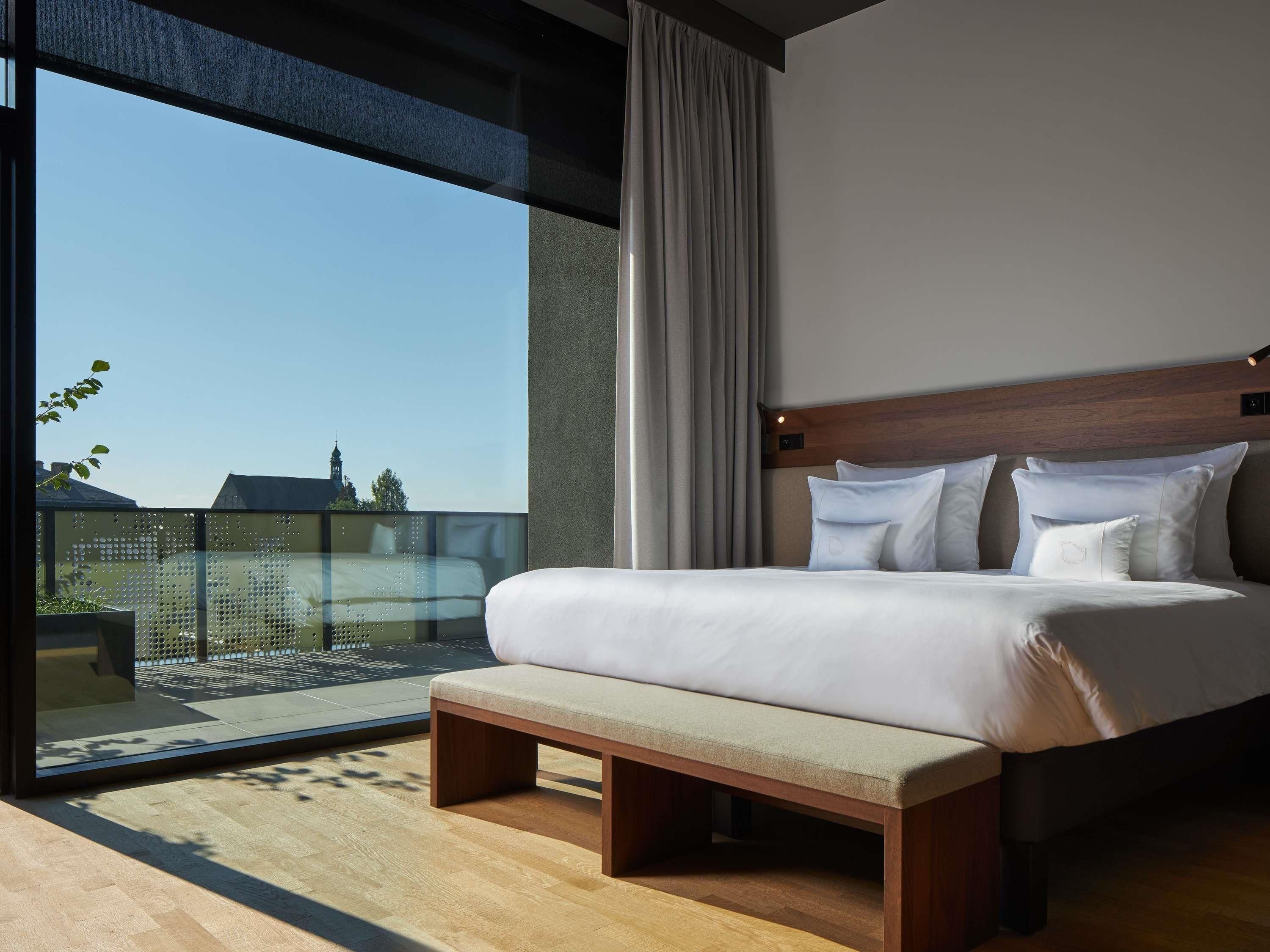 A modern bedroom with a large bed, a bench, a view of the sky, and a window.
